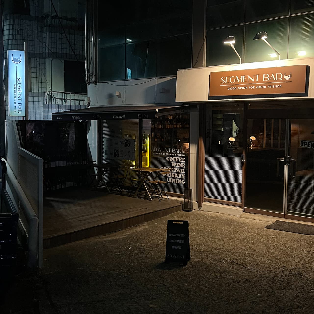 어두운 밤 조명이 비치는 건물 외관과 나무 데크 위 작은 테이블. 유리창 너머로 1인 이용이 가능한 바 좌석이 보이는 전경