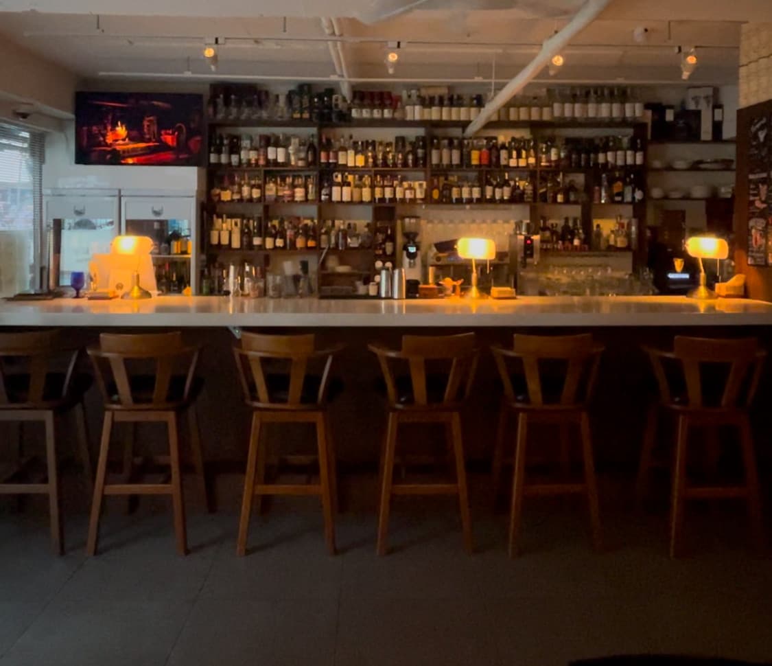 수많은 술병이 진열된 벽면 앞 긴 카운터와 목재 바 체어. 일정 간격으로 놓인 노란색 스탠드 조명이 비치는 일렬의 좌석들