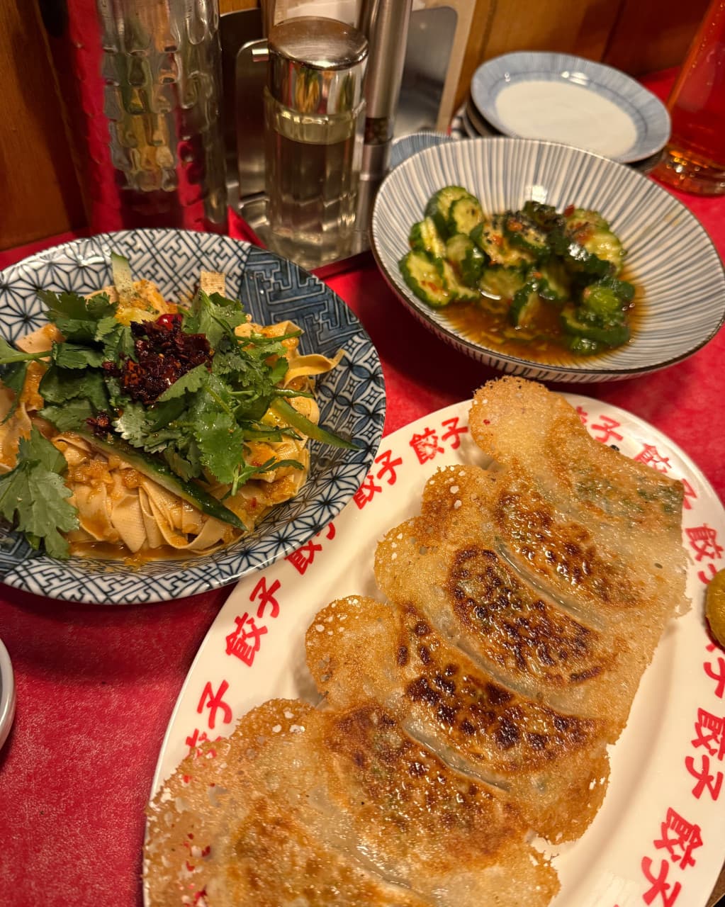 붉은색 바 테이블 위 바삭한 교자와 고수가 올라간 건두부 요리, 오이 무침이 나란히 놓인 1인용 안주 차림