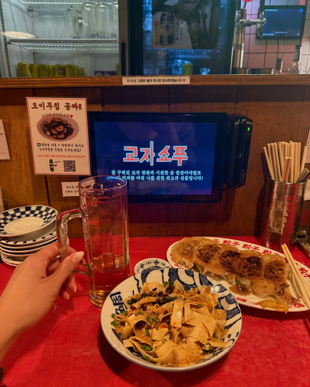 태블릿 메뉴판이 설치된 나무 다찌석 위 군만두와 면 요리, 하이볼 잔을 든 한 사람의 손