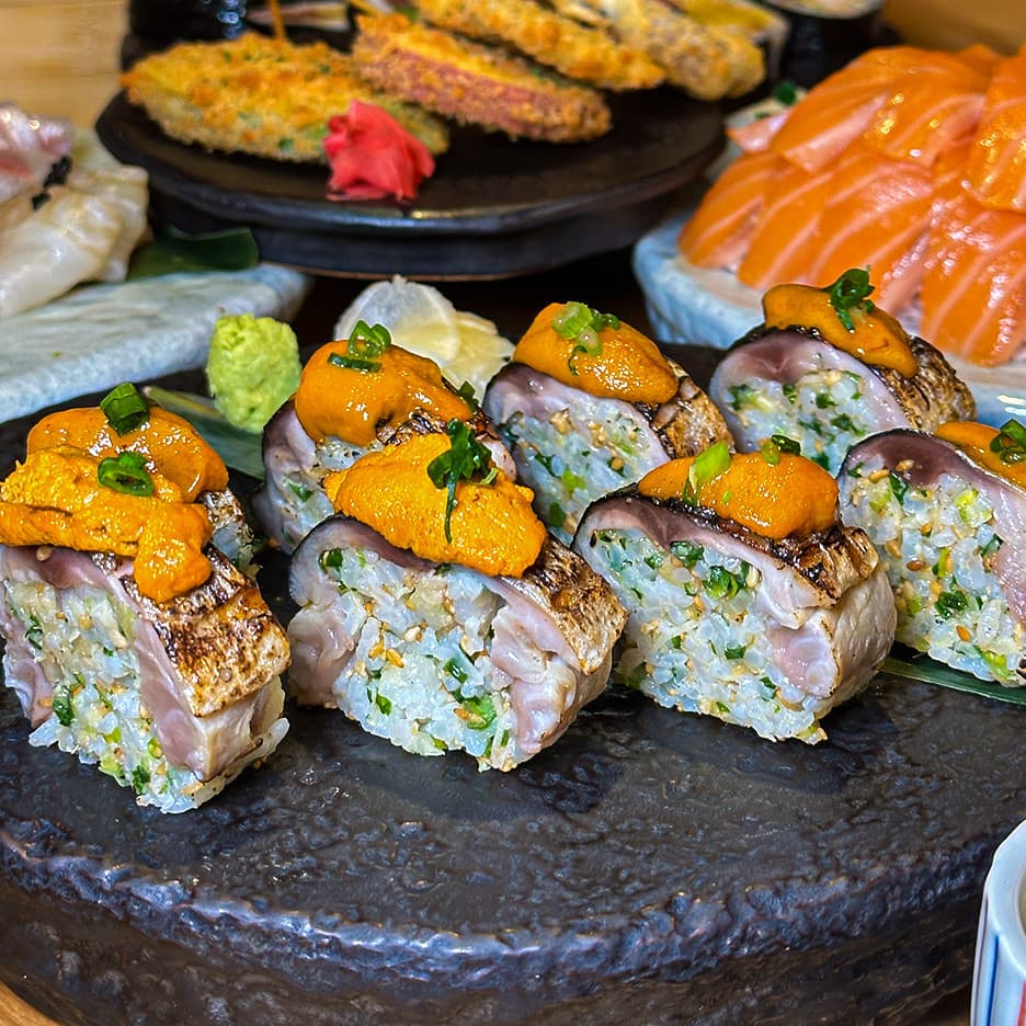 거친 질감의 검은 접시 위 성게알을 얹은 고등어 봉초밥과 배경에 차려진 연어 사시미 및 모듬 튀김