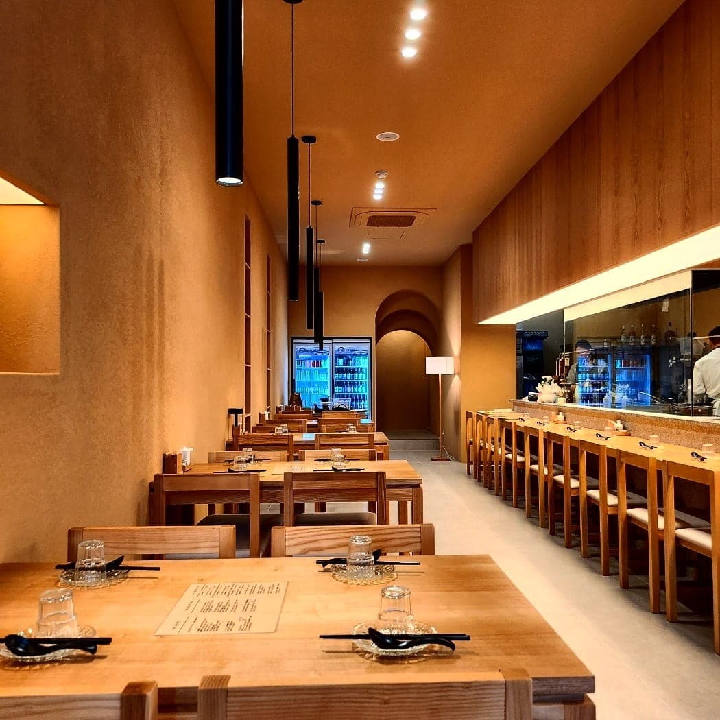 주황색 실내 벽면과 길게 이어진 나무 카운터석. 천장의 검은색 펜던트 조명 아래 1인용 식기가 세팅된 목재 테이블 전경