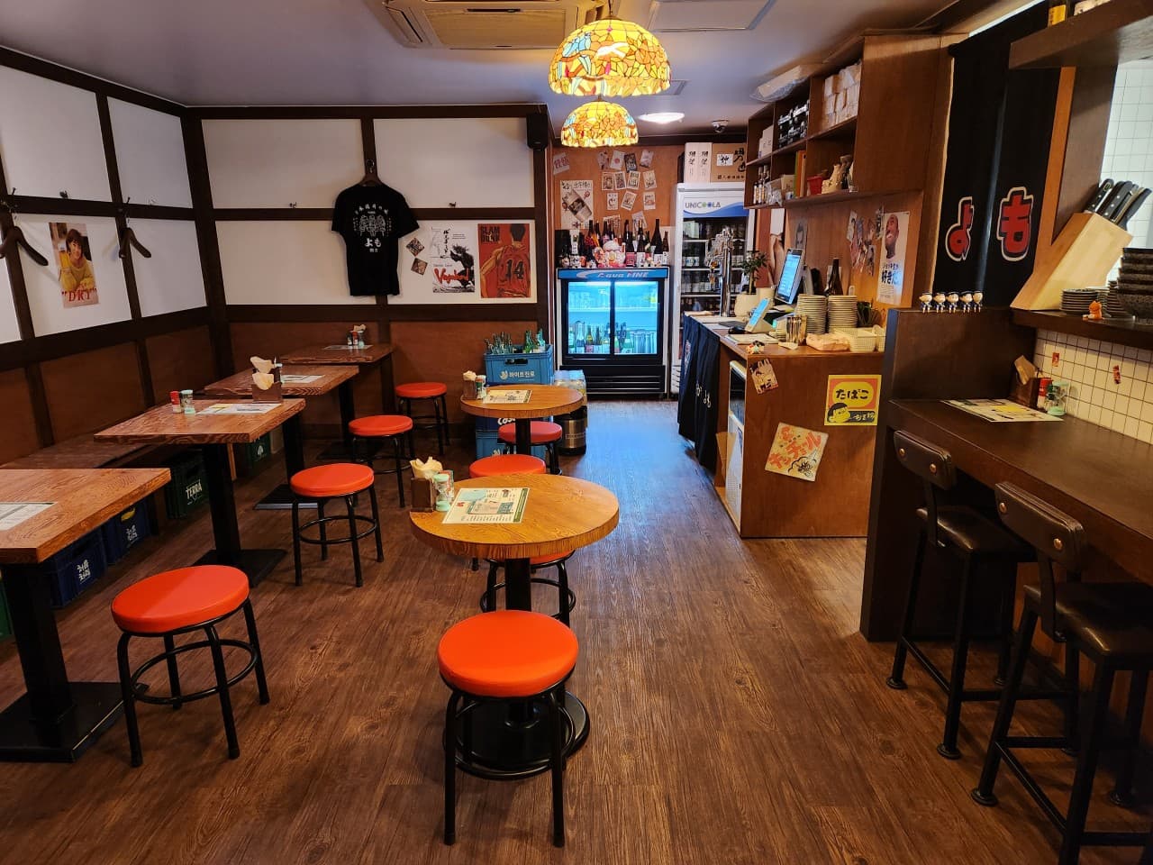 스테인드글라스 조명 아래 1인용 바 좌석과 소형 목재 테이블, 주황색 원형 의자가 배치된 실내