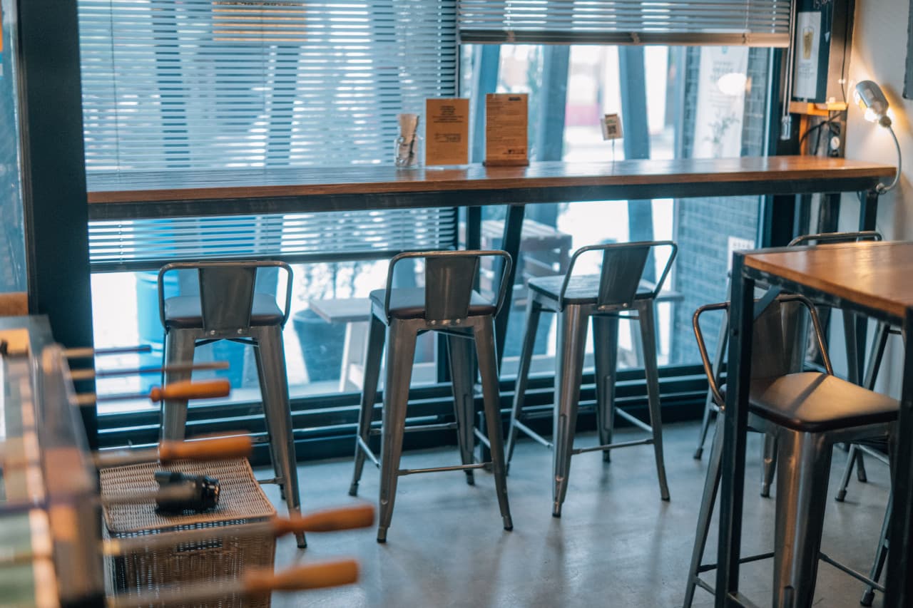 블라인드가 내려진 창가 쪽 긴 목재 카운터 테이블과 일렬로 배치된 높은 철제 바 스툴이 있는 실내 전경