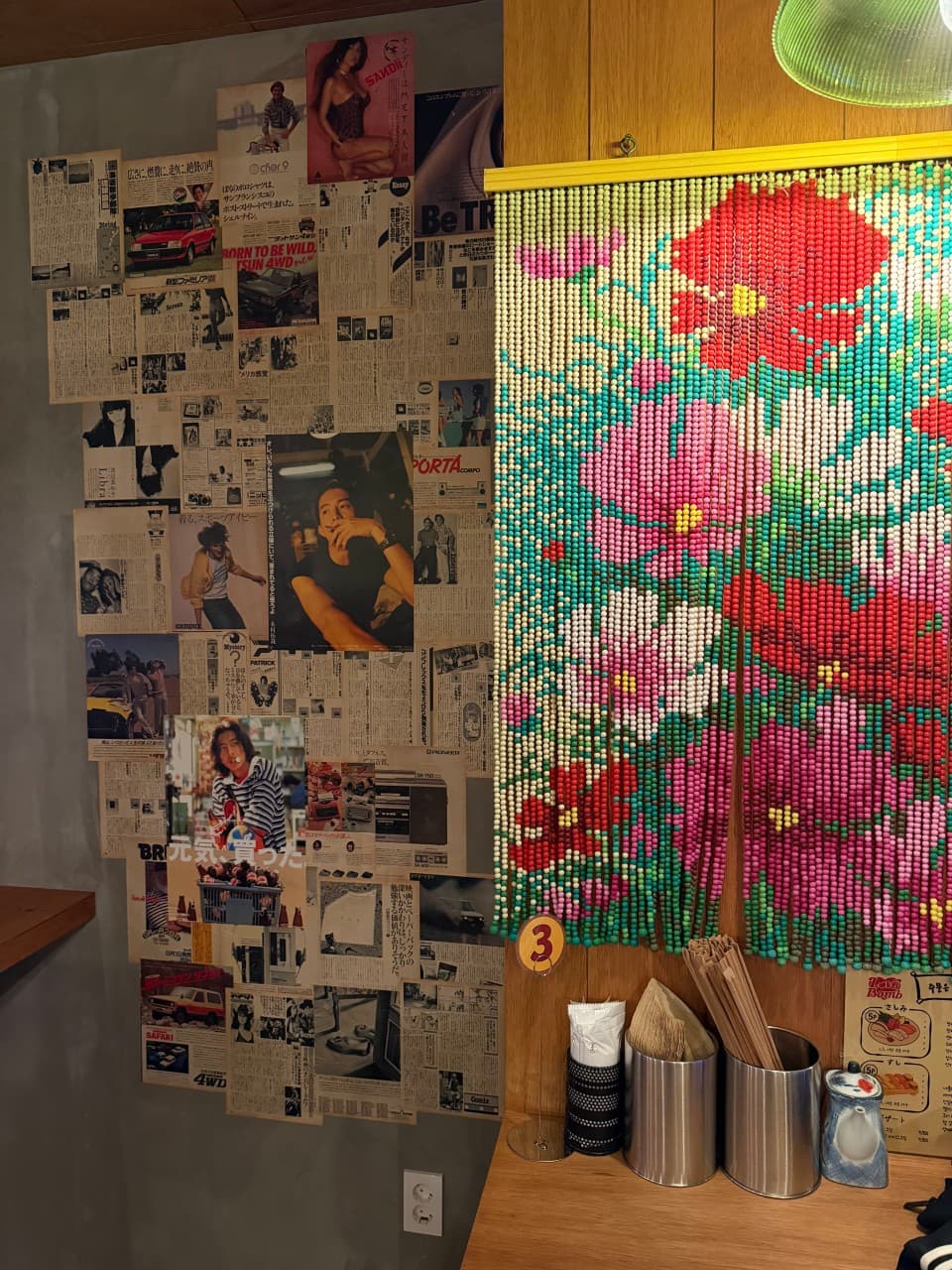 일본어 포스터와 광고가 붙은 벽면 옆 꽃무늬 구슬 발과 젓가락통이 놓인 1인용 목재 카운터