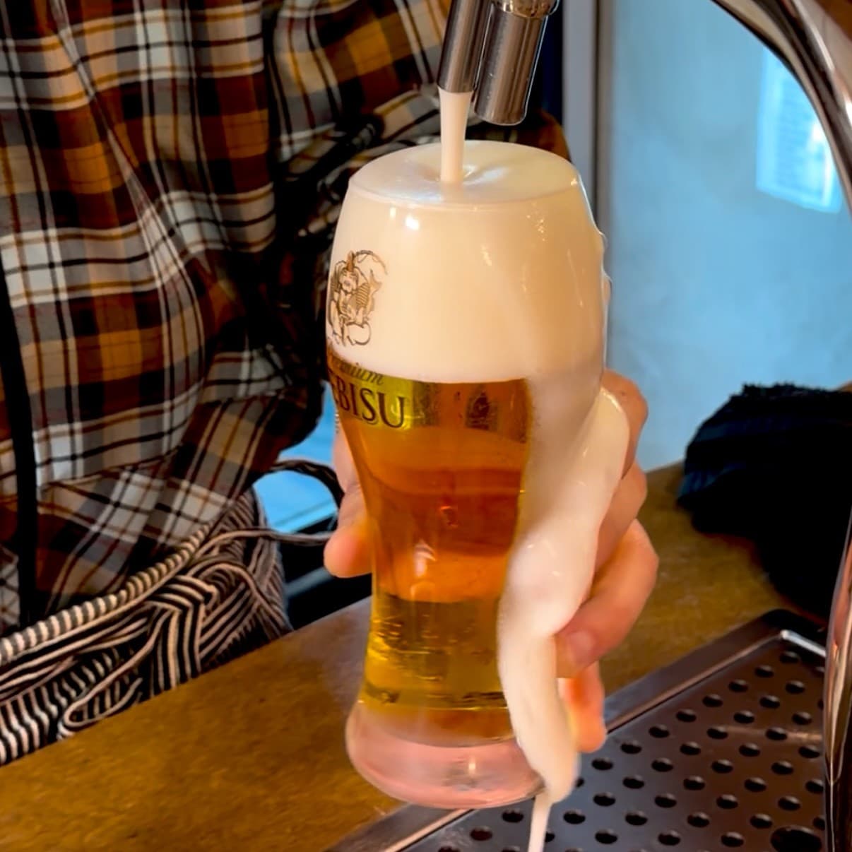 맥주 탭 아래 전용 잔에 가득 담긴 생맥주와 잔 옆으로 넘쳐흐르는 하얀 거품. 목재 카운터 위에 놓인 맥주잔과 금속 재질의 배수판.