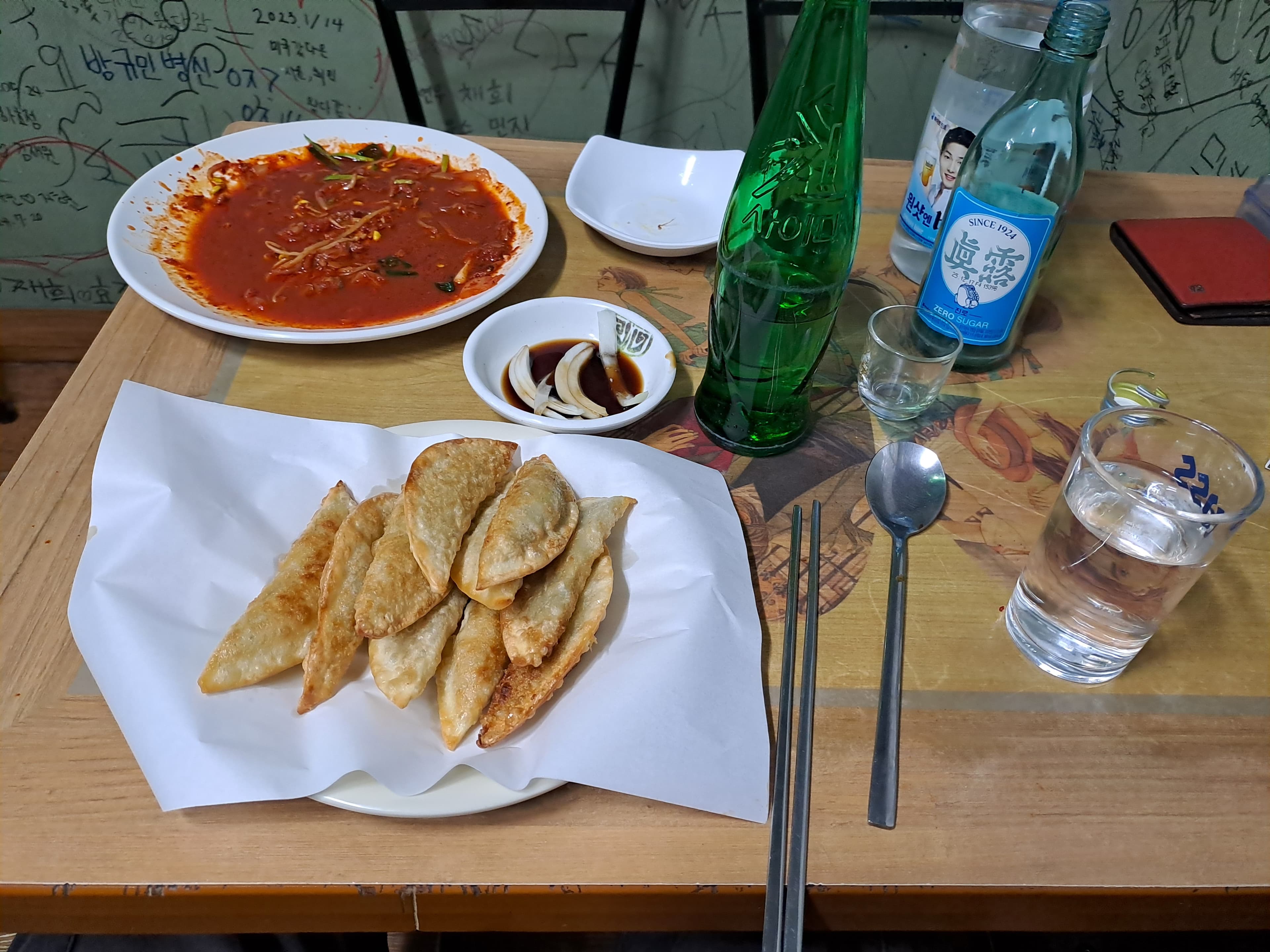 낙서가 있는 벽면 앞 나무 식탁 위 군만두와 붉은 소스 요리, 소주병과 초록색 유리병이 놓인 1인 상차림
