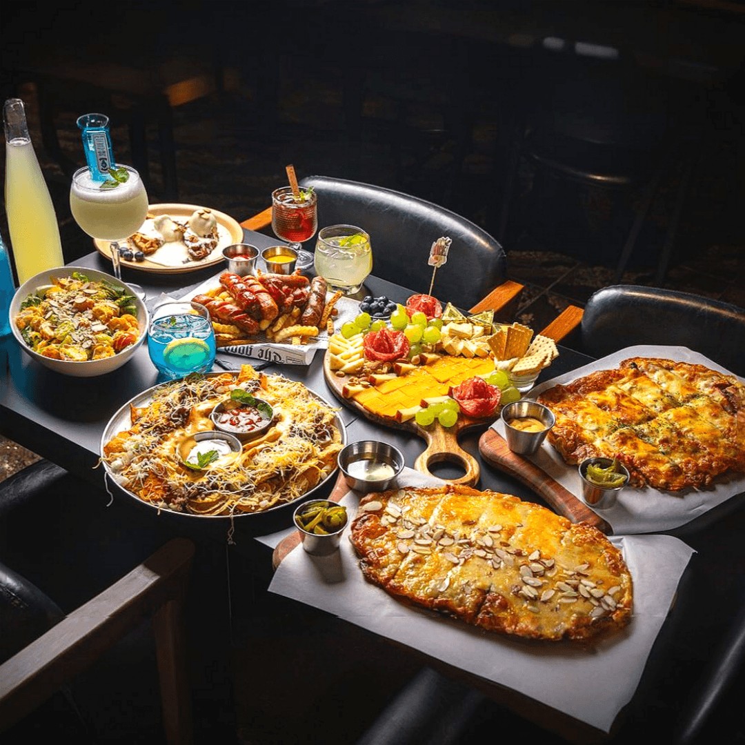 어두운 조명 아래 검은 테이블 위 치즈 피자와 나초, 소시지 플래터, 치즈 보드와 여러 색상의 하이볼 및 칵테일 잔