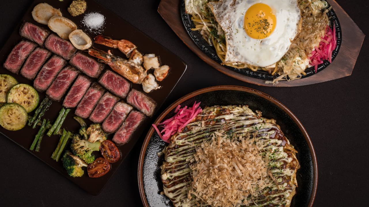 검은 테이블 위 소고기 스테이크와 구운 채소, 가쓰오부시가 뿌려진 오코노미야키와 계란 프라이를 얹은 야키소바 메뉴 구성