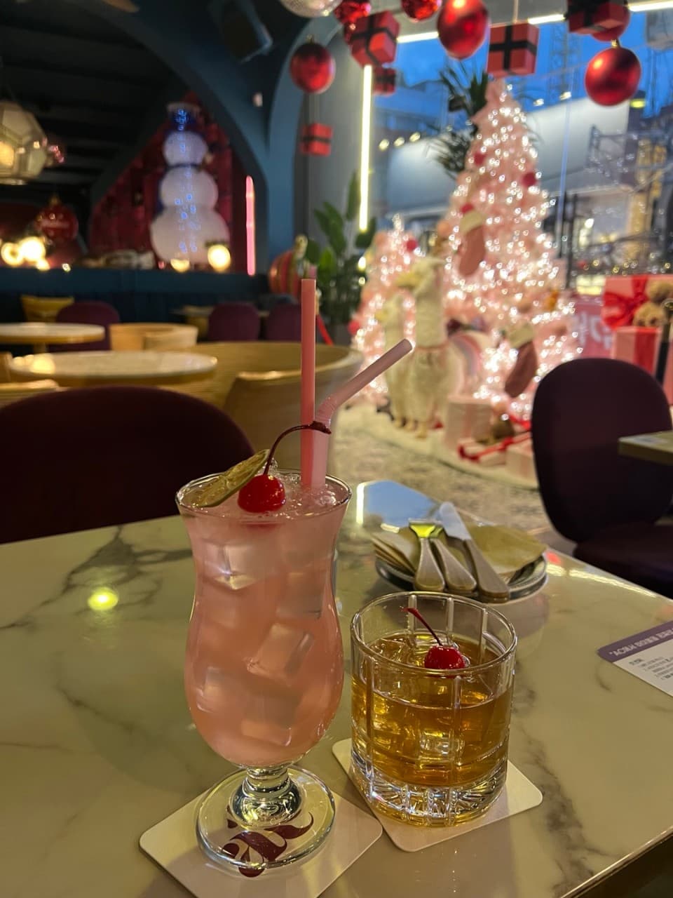 대리석 테이블 위 체리가 얹어진 분홍색 칵테일과 온더락 잔, 배경에 배치된 핑크빛 크리스마스 트리와 보라색 좌석들