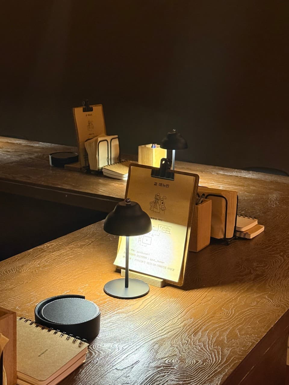 어두운 실내 나무 카운터 테이블 위 개별 조명과 클립보드 메뉴판, 메모용 수첩이 놓인 1인석