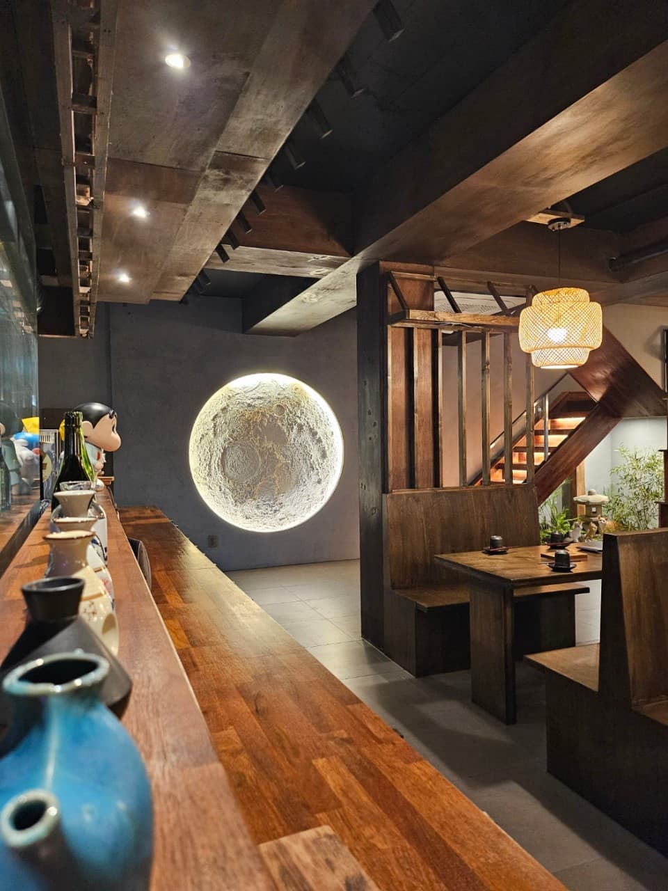 도자기 병들이 놓인 긴 나무 카운터석과 벽면의 커다란 원형 달 조명 및 목재 테이블이 배치된 공간