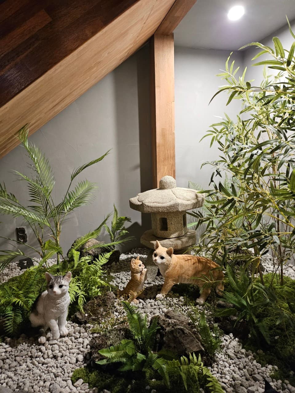 실내 계단 아래 흰 자갈과 이끼가 깔린 공간. 고양이 조형물과 석등, 대나무 조화가 배치된 정적인 조경 인테리어
