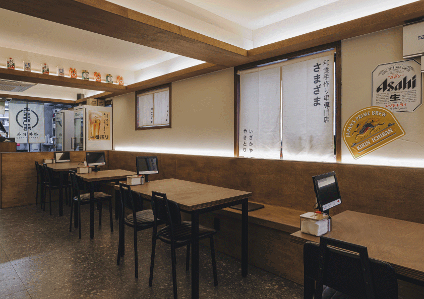 간접 조명이 비치는 나무 벽면 앞 2인용 테이블과 주문용 태블릿, 벽면에 부착된 일본풍 가림막과 맥주 광고 포스터