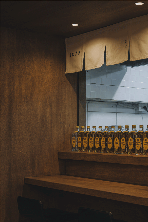 어두운 조명 아래 짙은 갈색 목재 카운터와 1인용 좌석 위 선반에 나란히 진열된 위스키 병들과 상단의 천 가림막