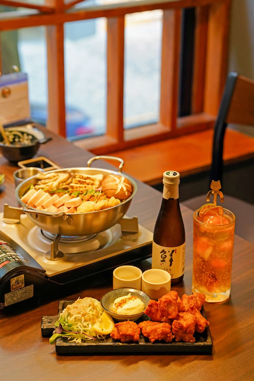 창가 나무 테이블 위 전골 냄비와 가라아게 접시, 사케 병과 고양이 장식 하이볼 잔이 놓인 상차림