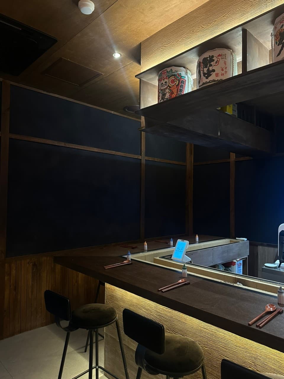 노란 조명이 비치는 목재 카운터에 놓인 1인용 수저 세트와 높은 의자. 상단 선반에는 사케 통이 진열된 어두운 이자카야 내부