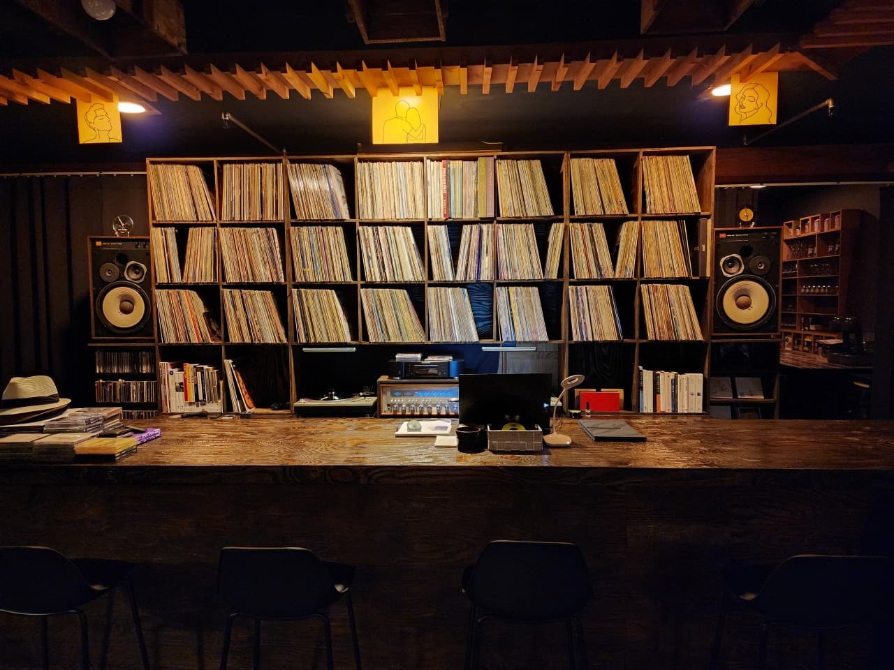 어두운 실내 속 대형 스피커와 LP판이 진열된 벽면을 마주 보는 긴 목재 카운터와 나란히 배치된 검은색 의자들