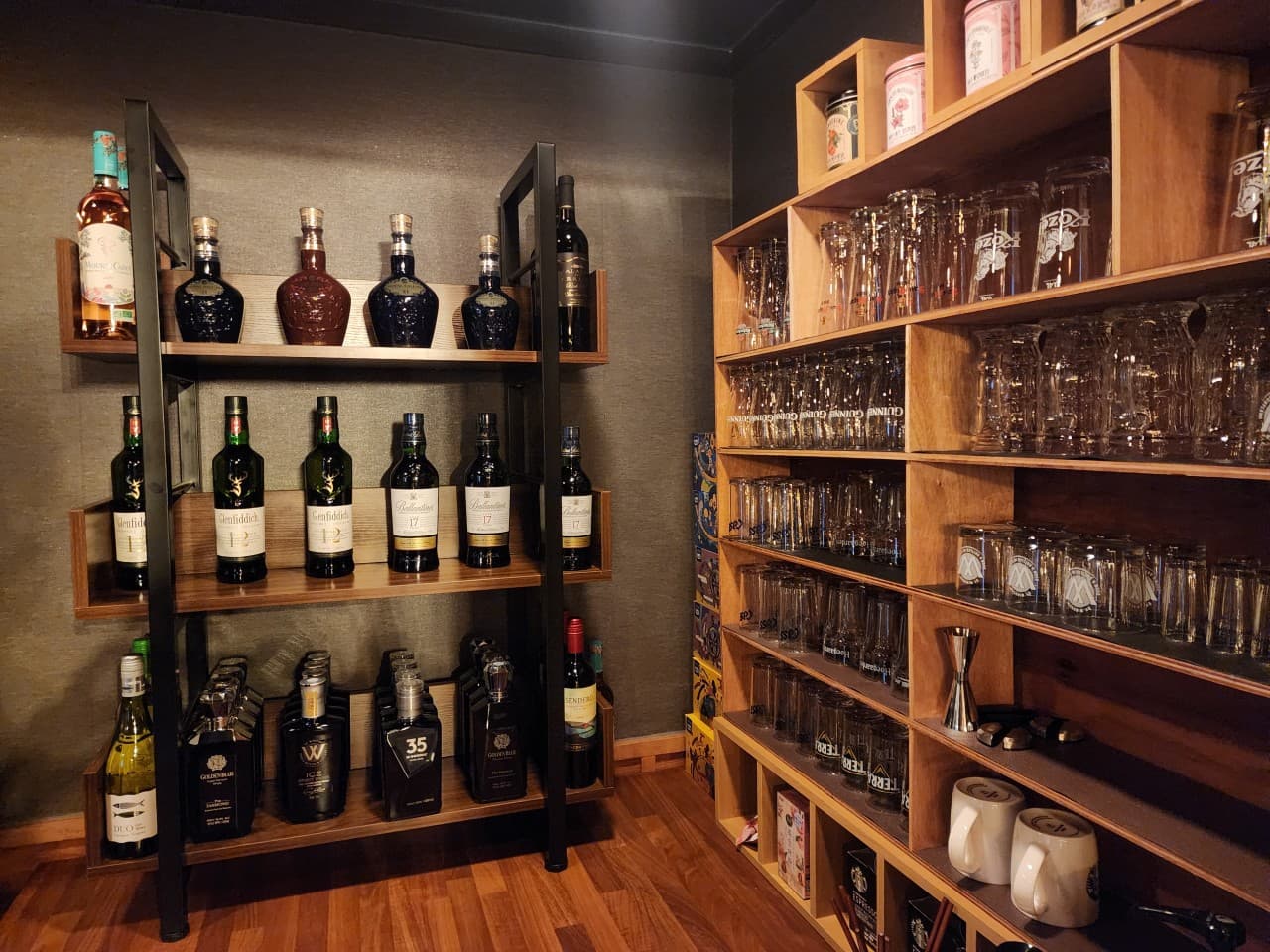 벽면 철제 선반에 진열된 위스키와 와인병, 그리고 옆면 목재 수납장을 가득 채운 맥주잔과 유리컵