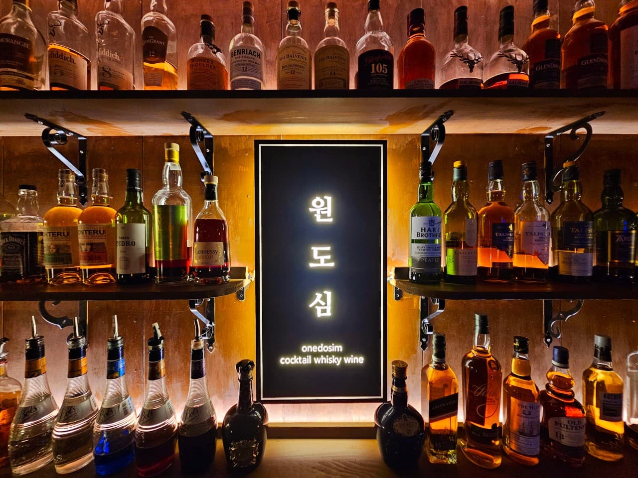 어두운 실내 속 노란 조명이 비치는 목재 선반 위 촘촘히 진열된 위스키 술병들과 중앙의 발광 사인