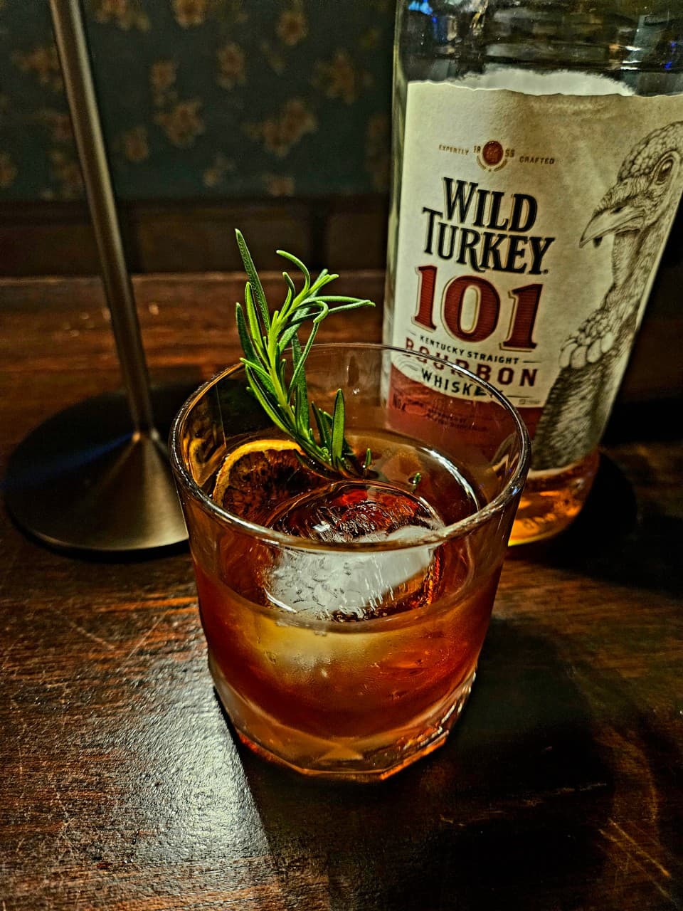 어두운 조명 아래 나무 질감의 테이블 위 로즈마리와 둥근 얼음이 담긴 위스키 잔과 버번 위스키 병