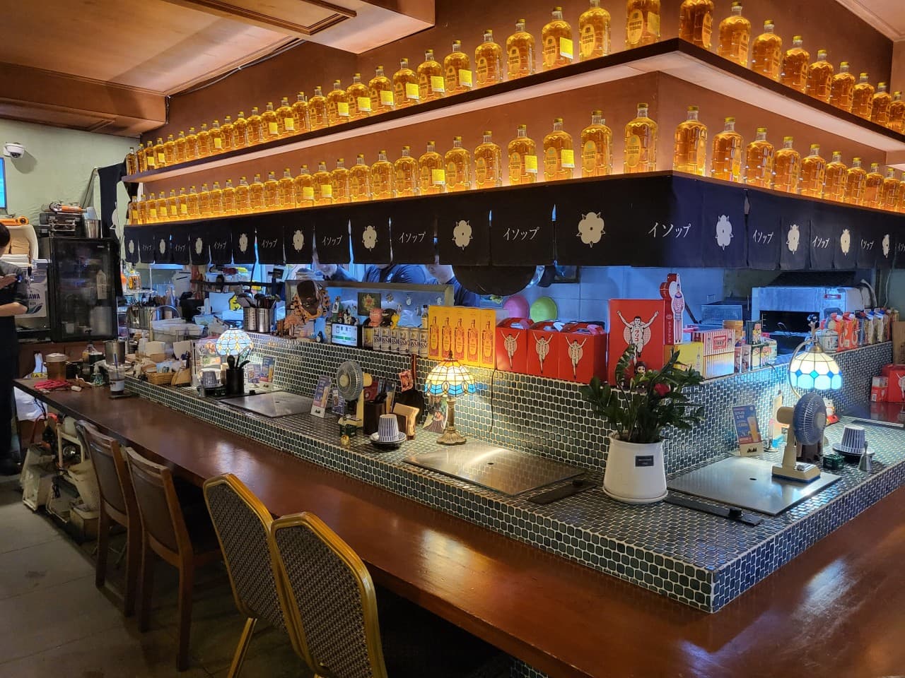 위스키 병이 진열된 선반 아래 타일로 마감된 카운터와 스테인드글라스 조명, 1인용 목재 의자가 배치된 바 테이블 전경
