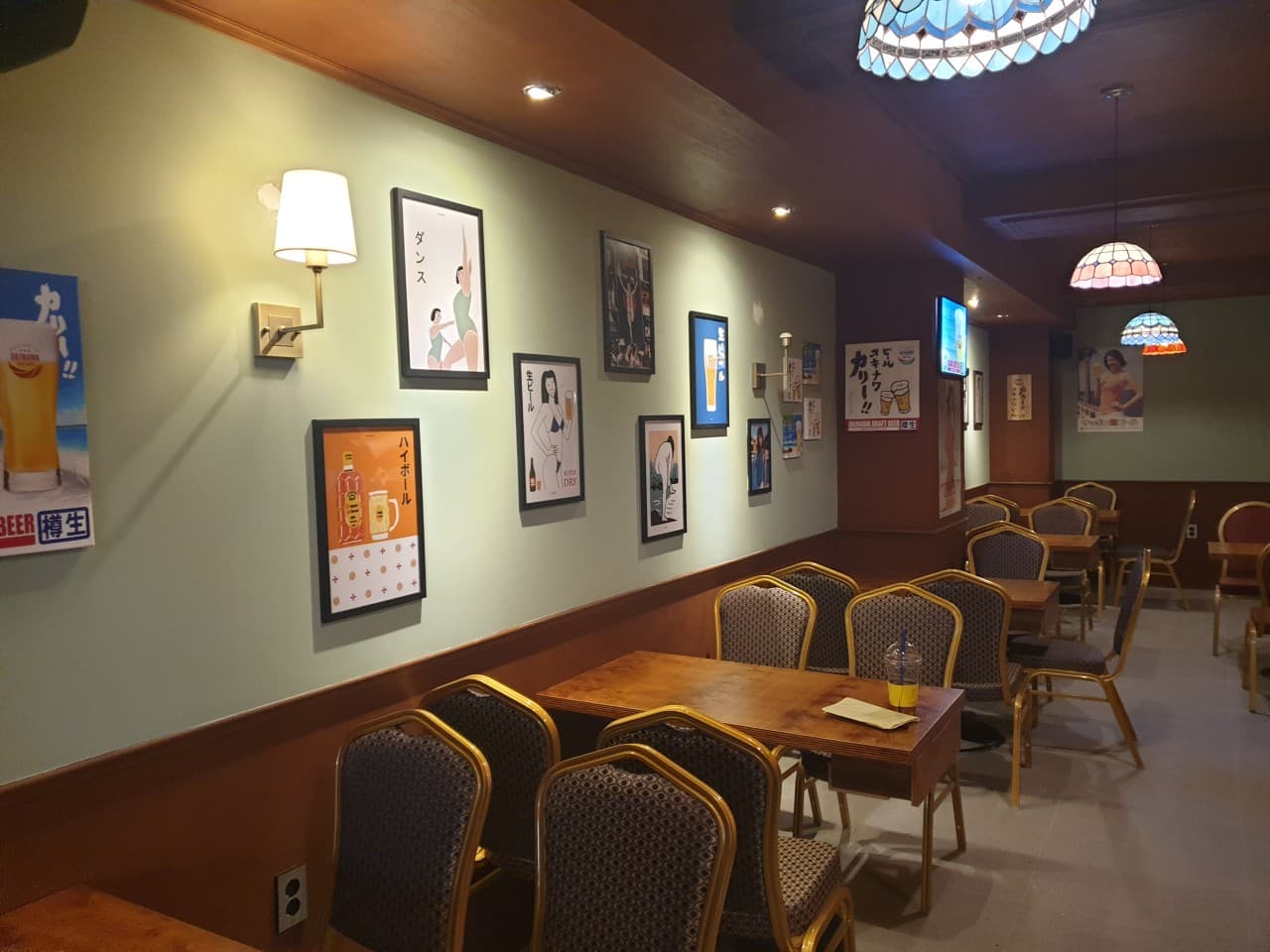 일본풍 포스터가 장식된 벽면과 주황색 펜던트 조명 아래 일렬로 배치된 소규모 목재 테이블과 의자