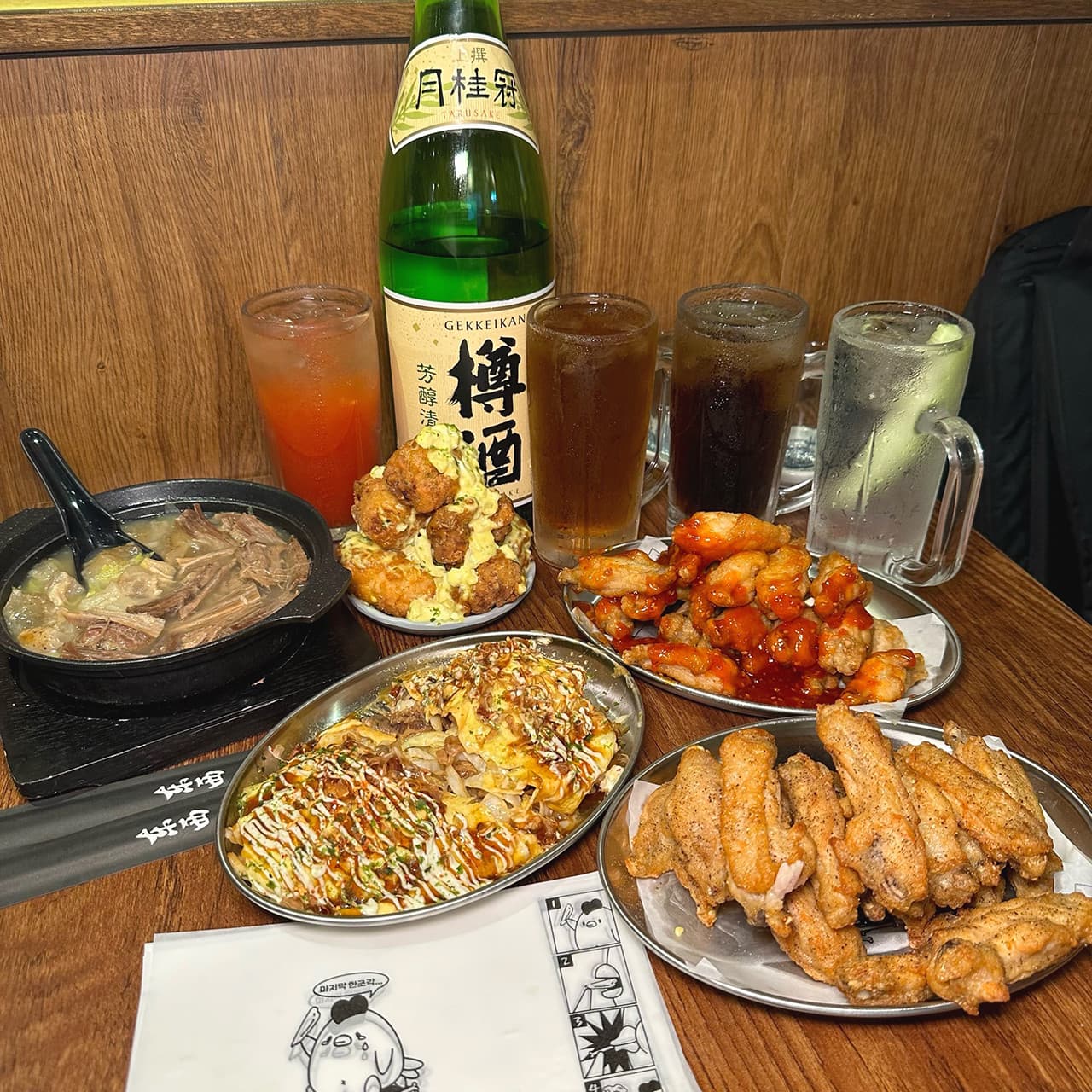 목재 테이블 위 사케 병과 하이볼 잔, 닭날개 튀김과 소스가 뿌려진 요리 등 작은 접시들로 구성된 일식 안주 상차림