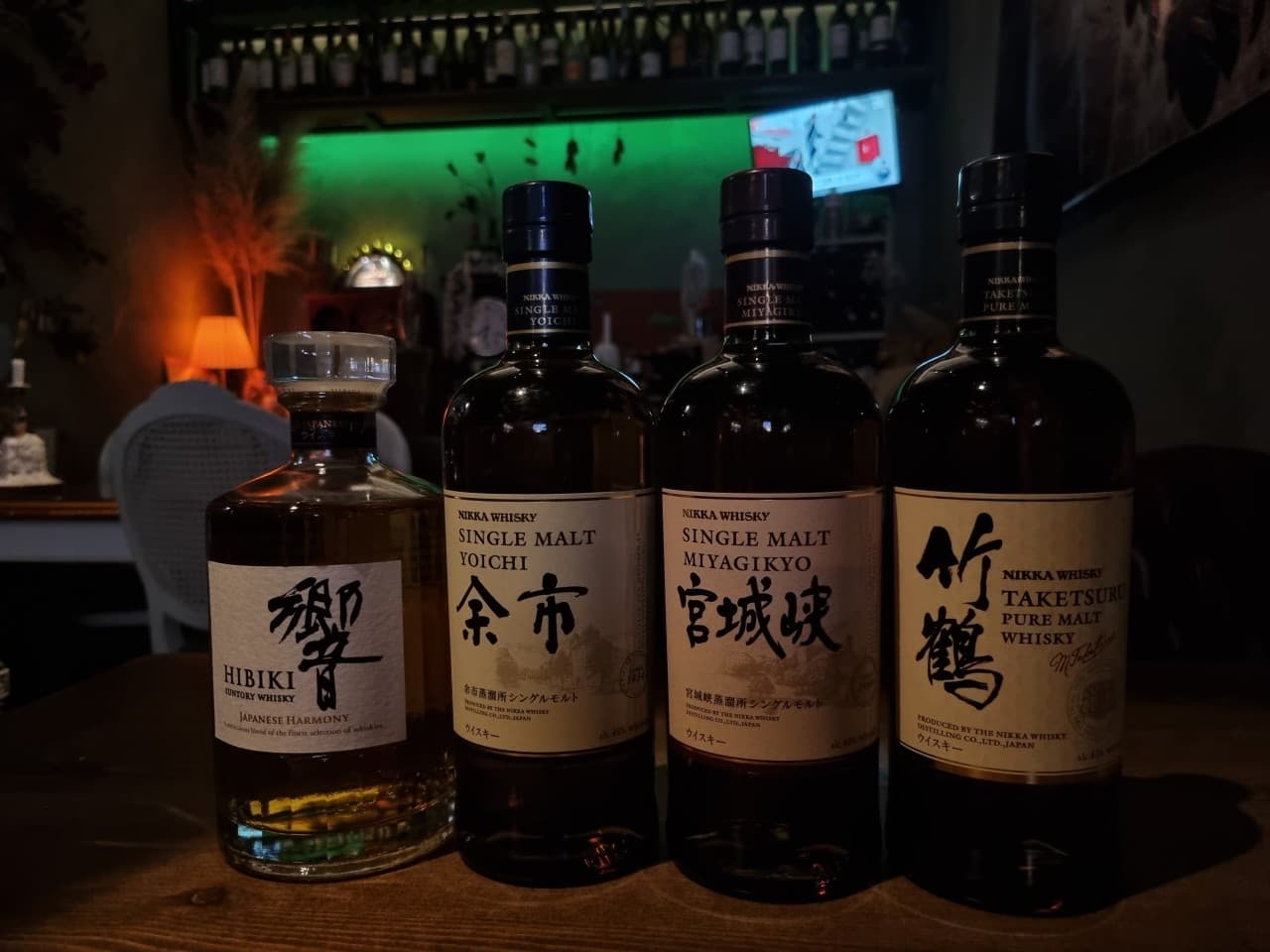 어두운 바 테이블 위 나란히 놓인 네 병의 위스키와 녹색 조명이 비치는 배경의 술 진열장