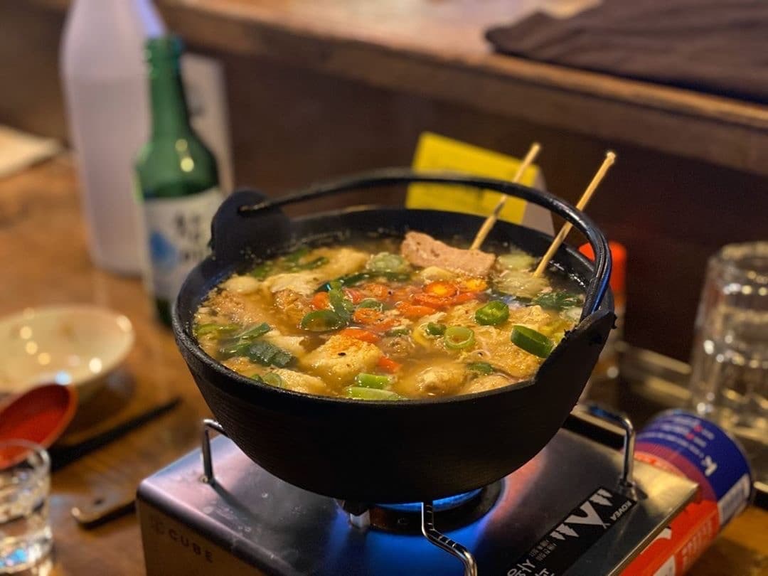 노란 조명이 비치는 나무 카운터 위 휴대용 가스레인지에서 끓는 어묵탕과 소주병