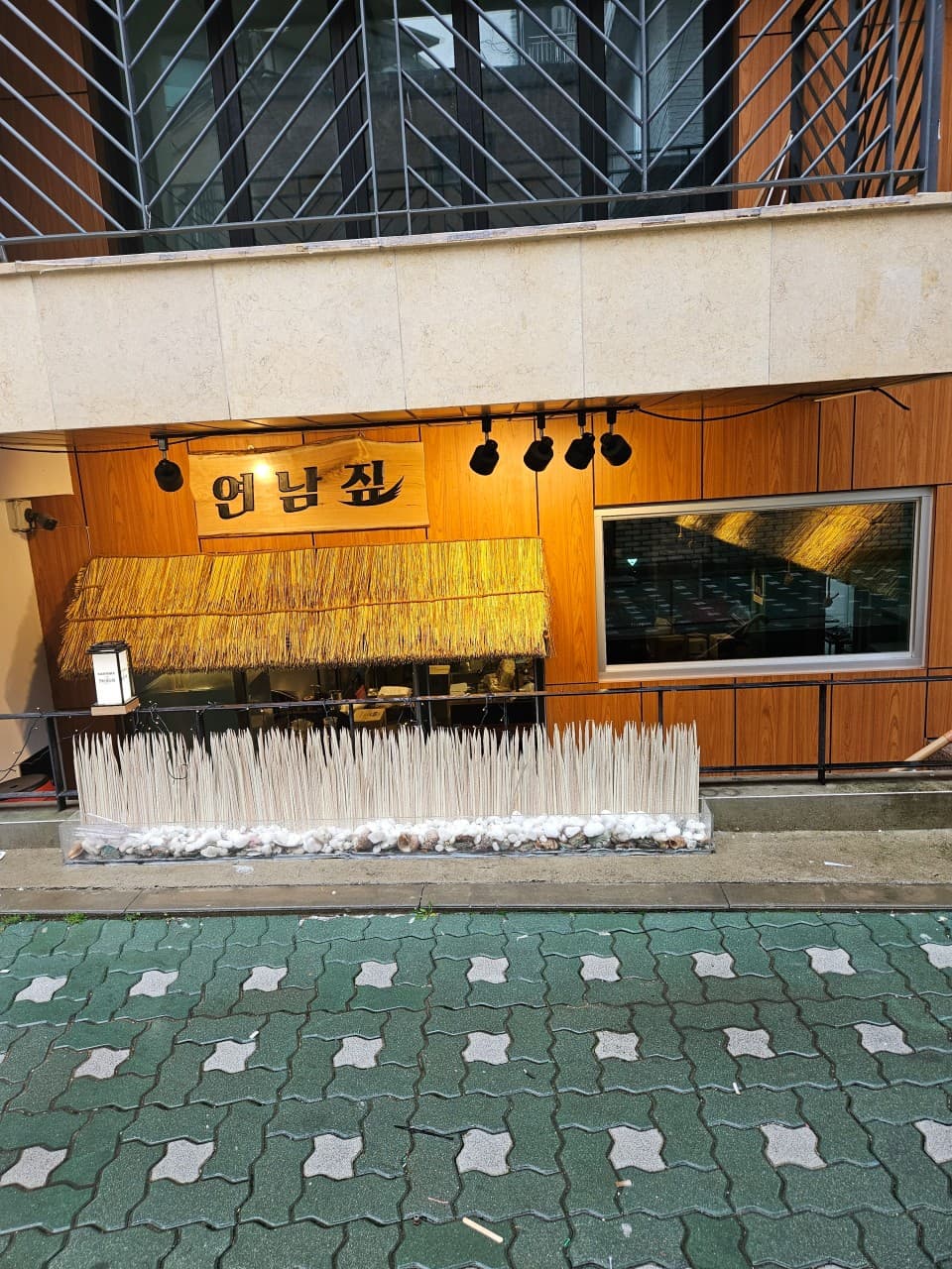 짚으로 된 지붕 장식과 목재 외벽, 검은색 조명이 설치된 입구와 하단에 배치된 흰색 자갈 및 수직 장식물