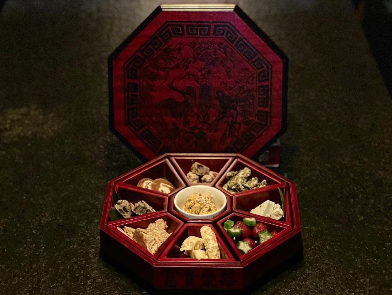 어두운 배경의 테이블 위 붉은색 팔각형 찬합과 칸마다 나누어 담긴 견과류, 부각, 말린 과일 등의 소량 안주