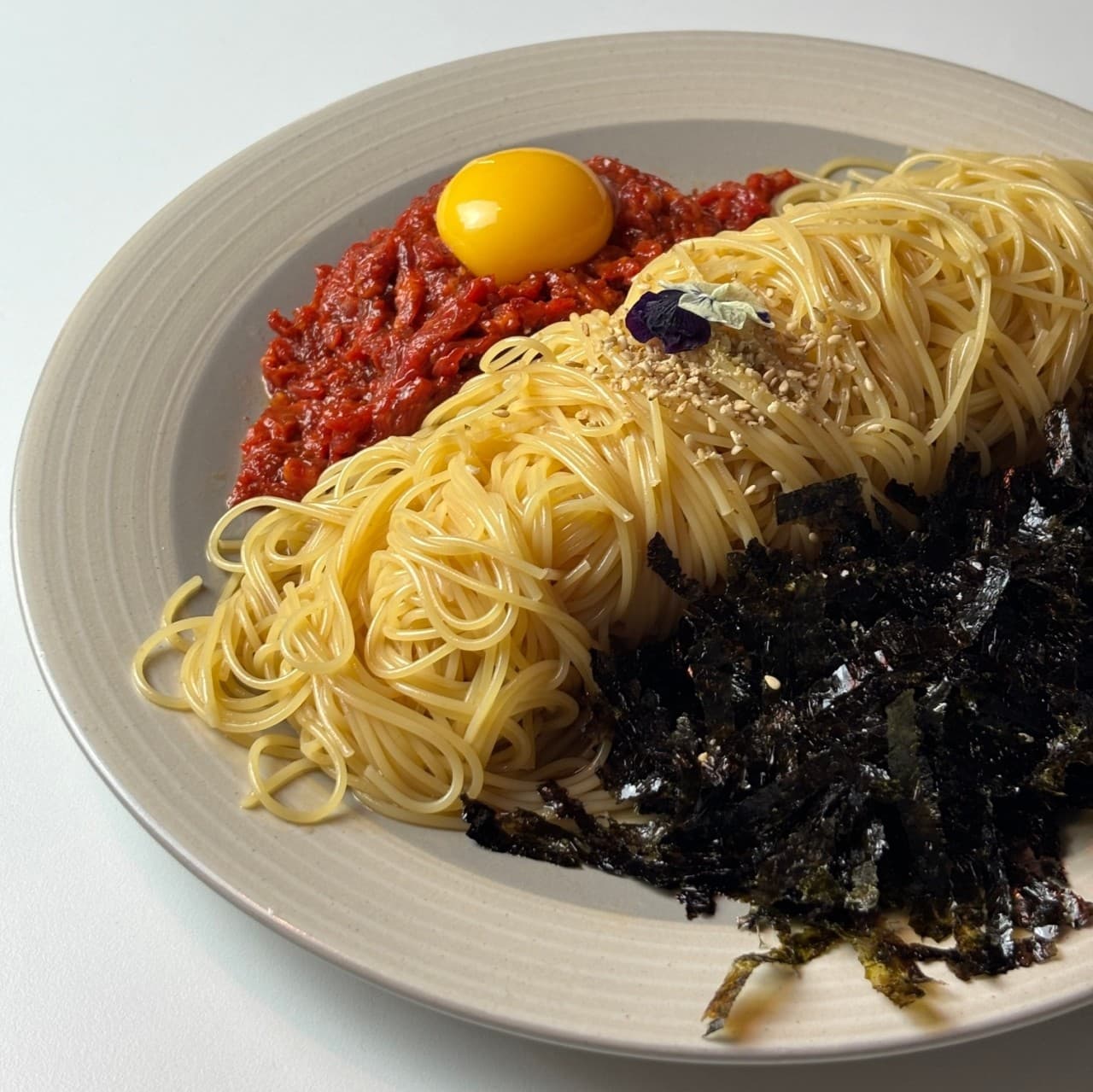 밝은 배경 위 1인용 접시에 담긴 얇은 면 파스타와 붉은 양념 고기, 달걀노른자와 김 가루가 곁들여진 차림