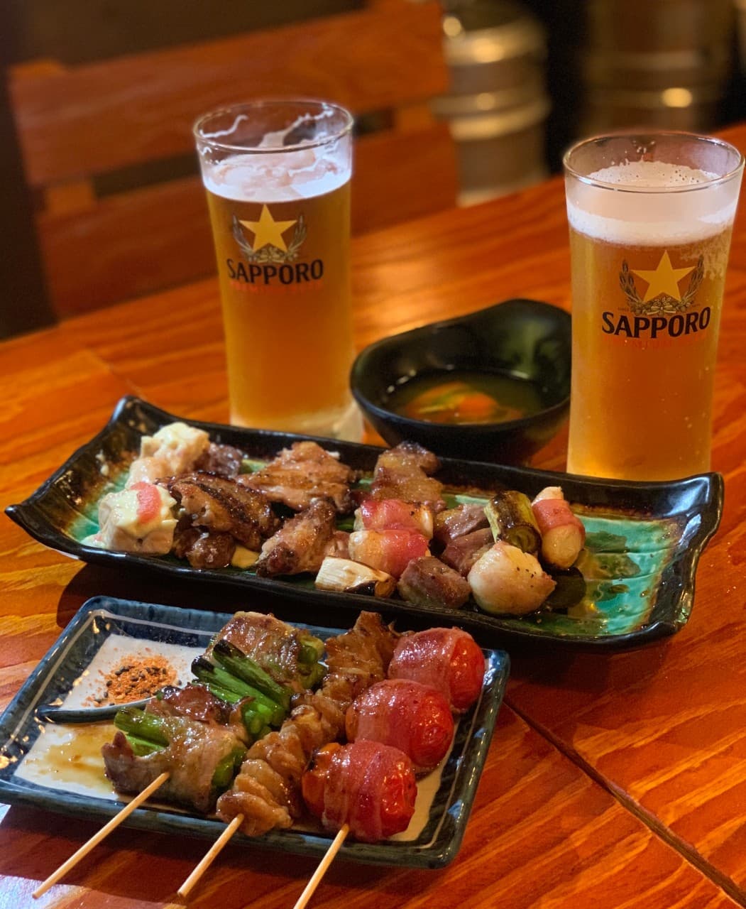 황색 조명이 비치는 나무 테이블 위 삿포로 맥주잔과 베이컨 토마토 말이, 대파 꼬치 등 모둠 꼬치 메뉴