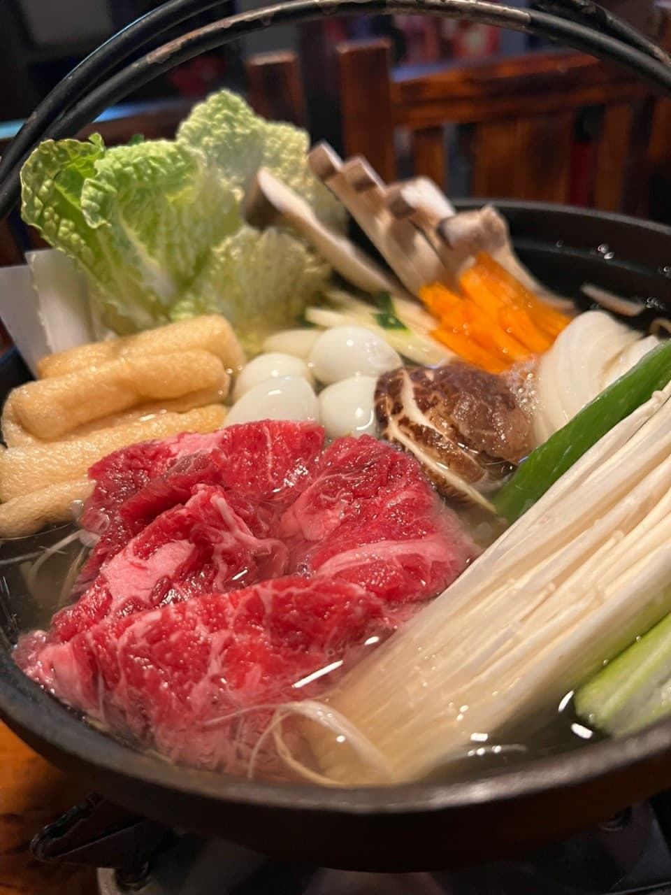 어두운 조명 아래 목재 테이블 위 무쇠 냄비에 담긴 선홍색 소고기와 팽이버섯, 유부, 메추리알 등 1인 구성의 전골