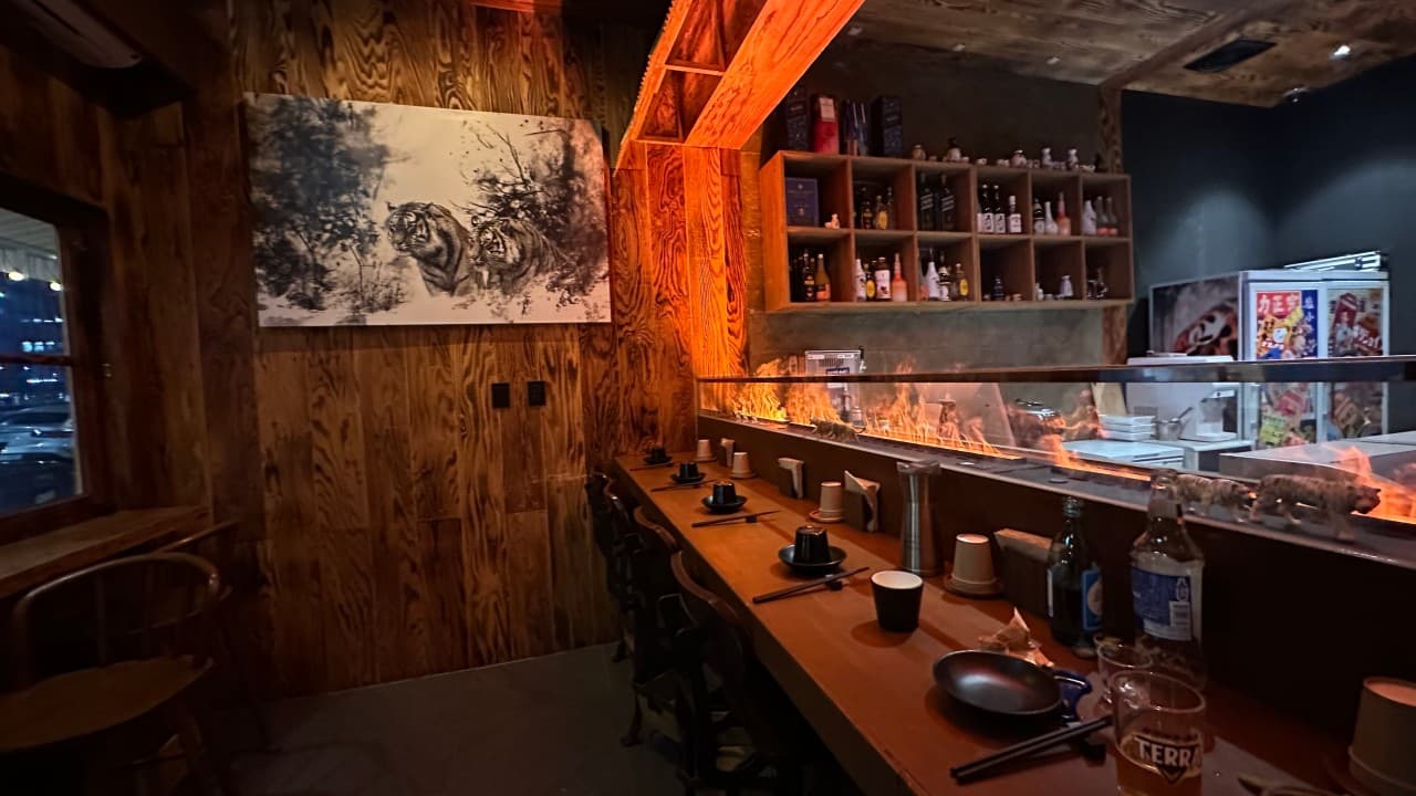 호랑이 그림이 걸린 목재 벽면 아래 주황색 조명이 비치는 바 테이블과 일정한 간격으로 배치된 1인용 식기