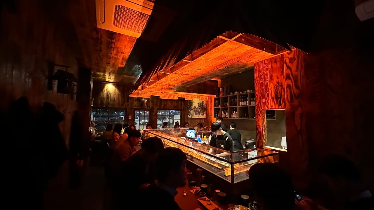 어두운 실내 속 주황색 조명이 비치는 긴 나무 카운터석과 일렬로 배치된 좌석, 오픈 주방에서 조리하는 직원과 선반 위 술병들