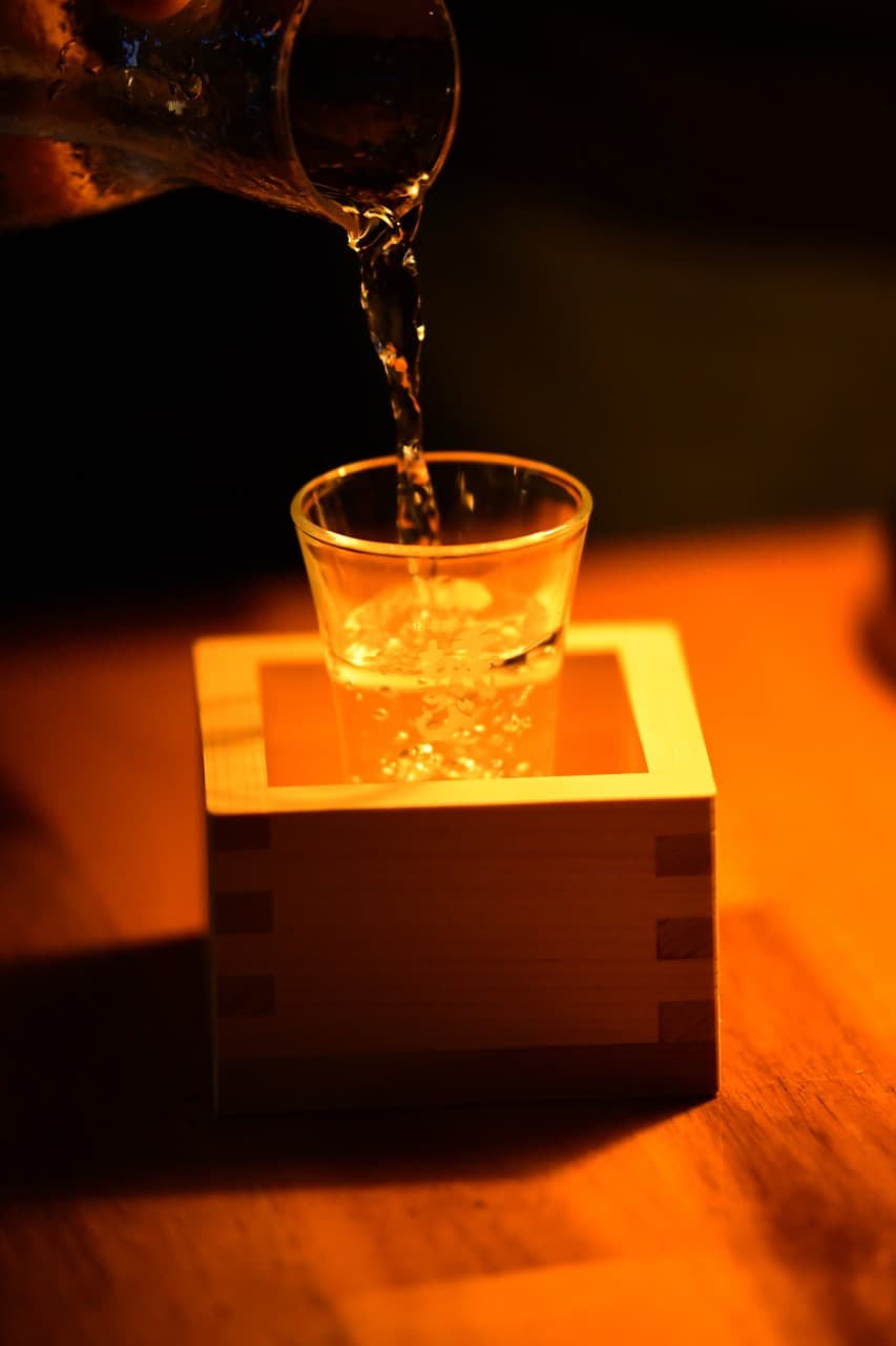 경기도 수원시 고래다 수원 장안 오뎅바 주황색 조명 아래 1인 다찌석에서 사케를 따르는 혼술 상황. 나무 상자 속 유리잔과 투명한 술의 질감이 돋보임.