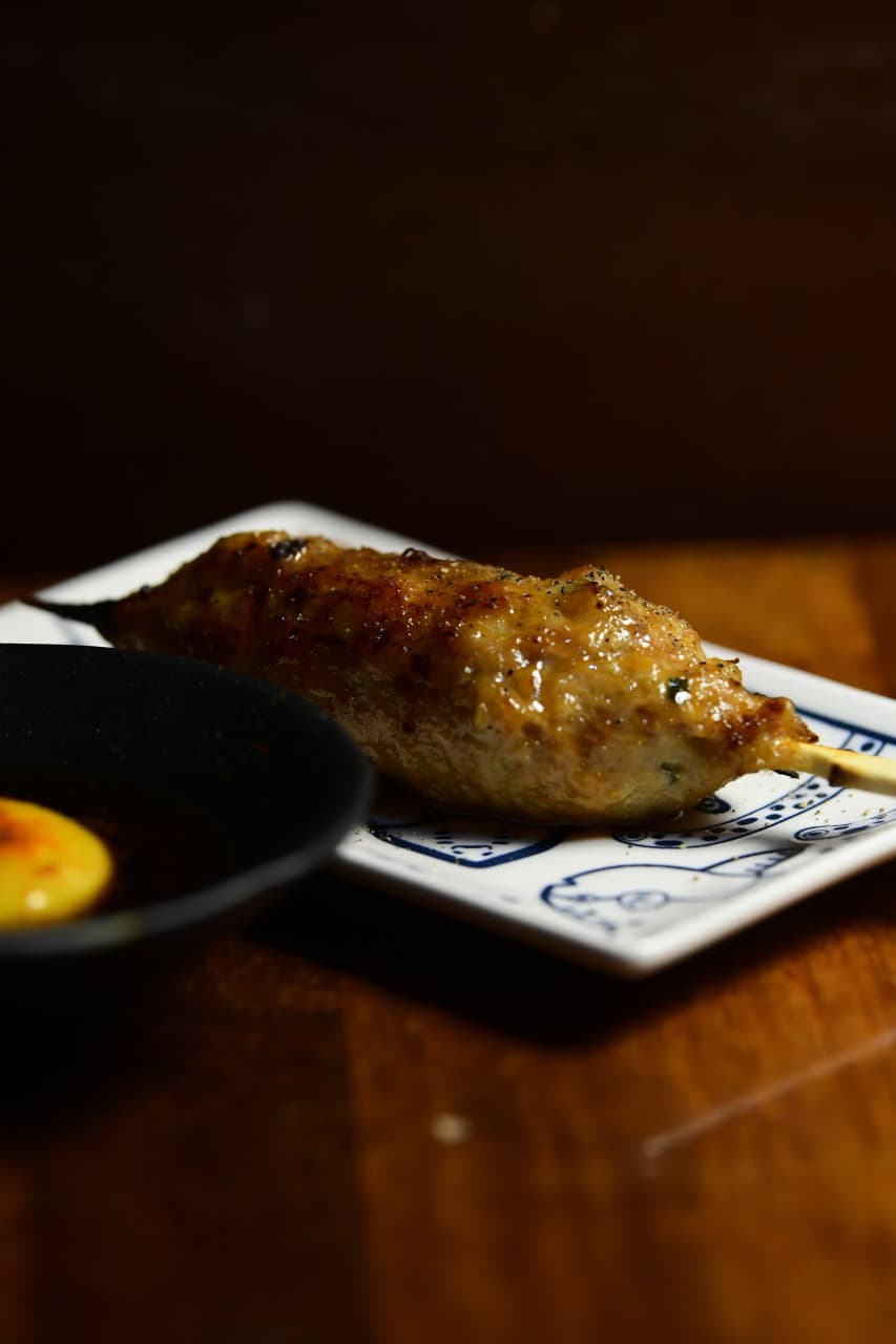 경기도 수원시 고래다 수원 장안 오뎅바의 어두운 조명 아래 나무 다찌석 1인 혼술 차림. 노른자 소스를 곁들인 윤기 나는 닭꼬치 메뉴.