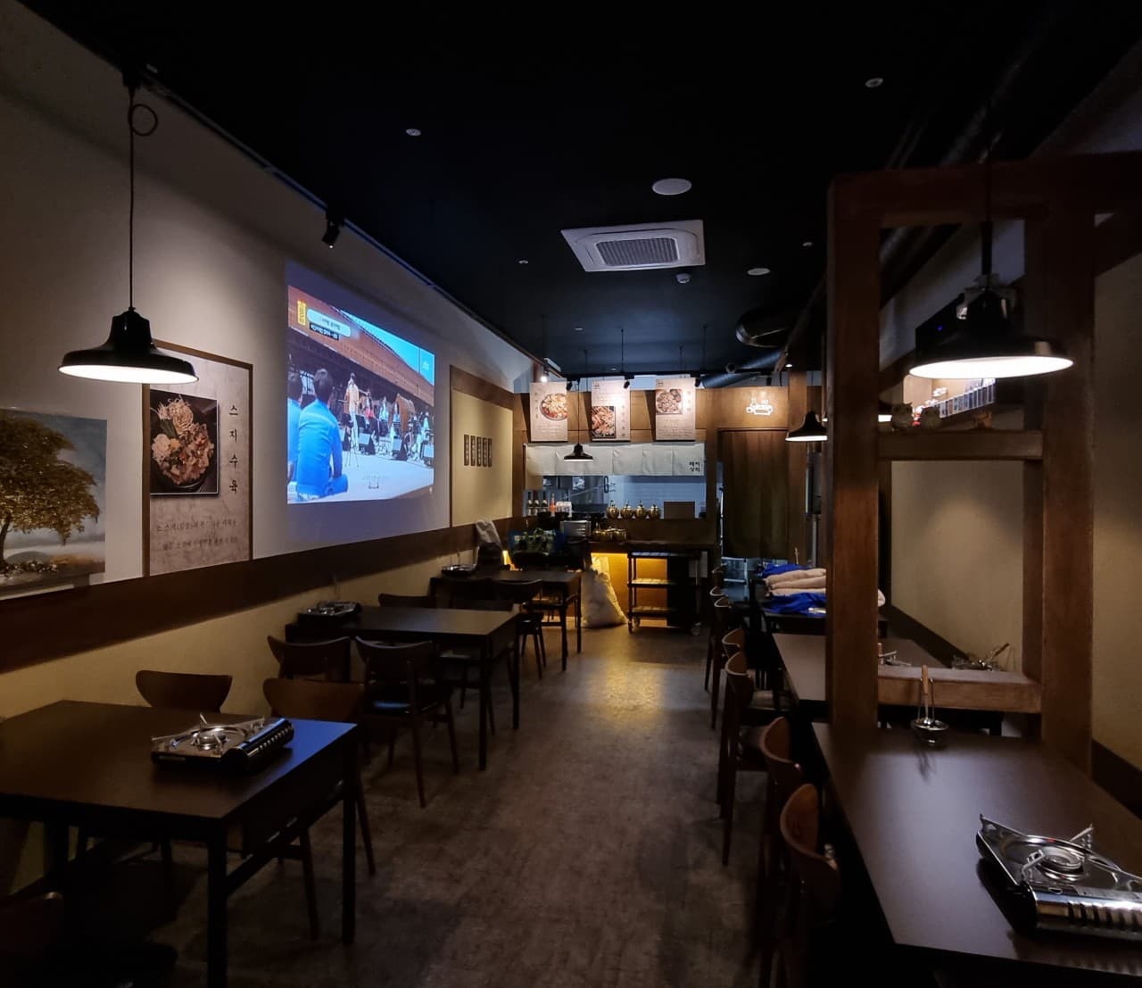 경기도 수원시 혼술 요리주점 배씨상회 수원스타필드 - 조명 아래 빔프로젝터 영상이 나오는 벽면과 1인용 다찌석 및 버너가 놓인 목재 테이블