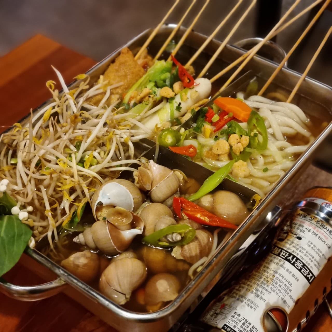 경기도 수원시 배씨상회 수원스타필드 요리주점 다찌석. 주황색 조명 아래 칸막이 냄비에 담긴 꼬치 어묵과 골뱅이, 우동, 숙주가 정갈하게 배치된 1인 혼술 차림.