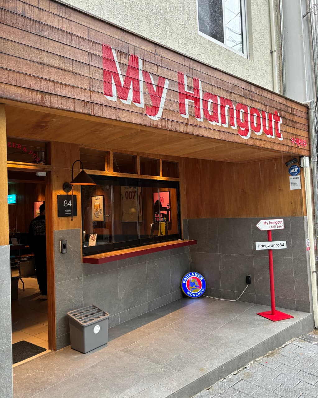 부산광역시 중구 마이행아웃 맥주 호프 - 우드 외벽과 붉은 간판 아래 다찌석이 보이는 1인 혼술 장소. 매장 밖 파울라너 조명과 내부의 노란 조명이 켜진 모습.