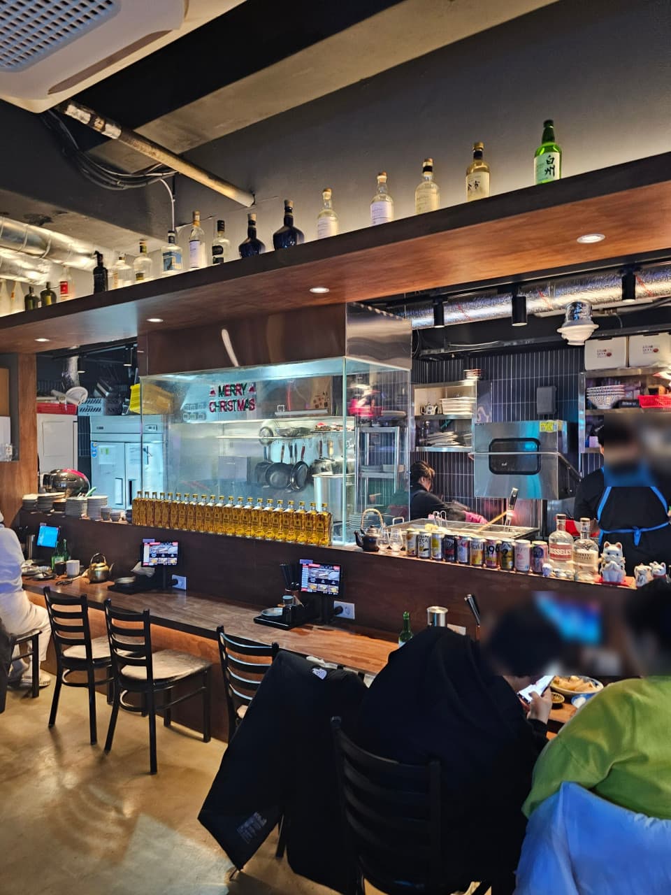 부산광역시 중구 요리주점 히노카오뎅 남포점 - 따뜻한 핀 조명 아래 우드 다찌석과 1인용 태블릿이 설치된 공간에서 술병이 진열된 선반을 마주하며 즐기는 혼술 환경