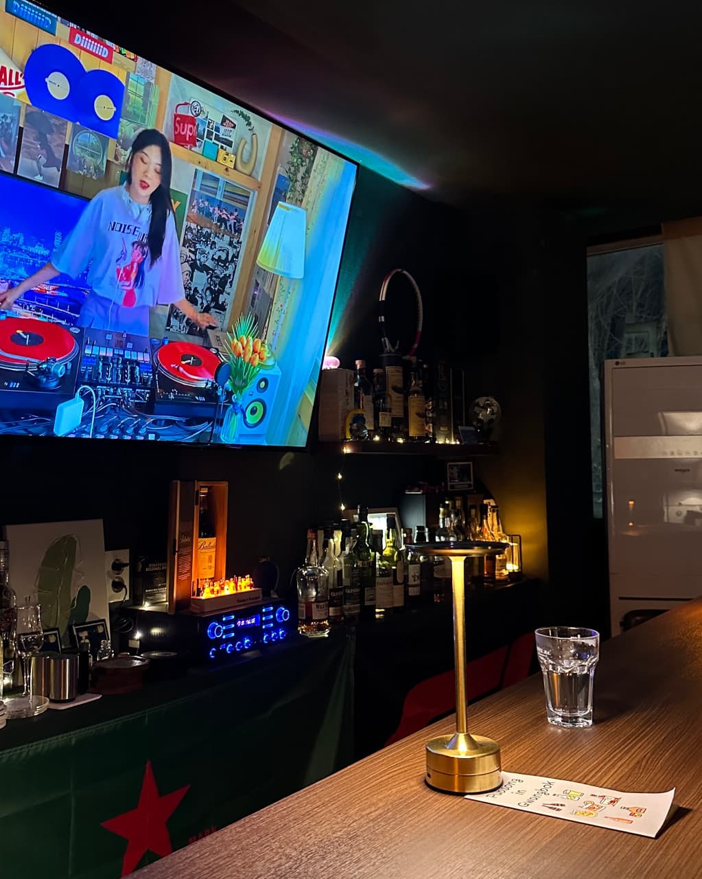 부산광역시 중구 맥주 호프 펍송 인 광복 - 대형 스크린 영상과 테이블 조명이 켜진 1인 다찌석, 여러 술병이 진열된 어두운 실내 혼술 공간