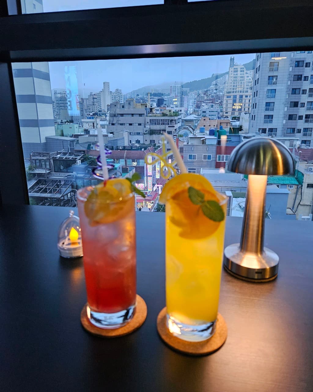 부산광역시 중구 혼술 바 선셋라운지 - 창가 1인 다찌석 시티뷰 배경으로 스탠드 조명과 함께 놓인 과일 가니시 칵테일 두 잔
