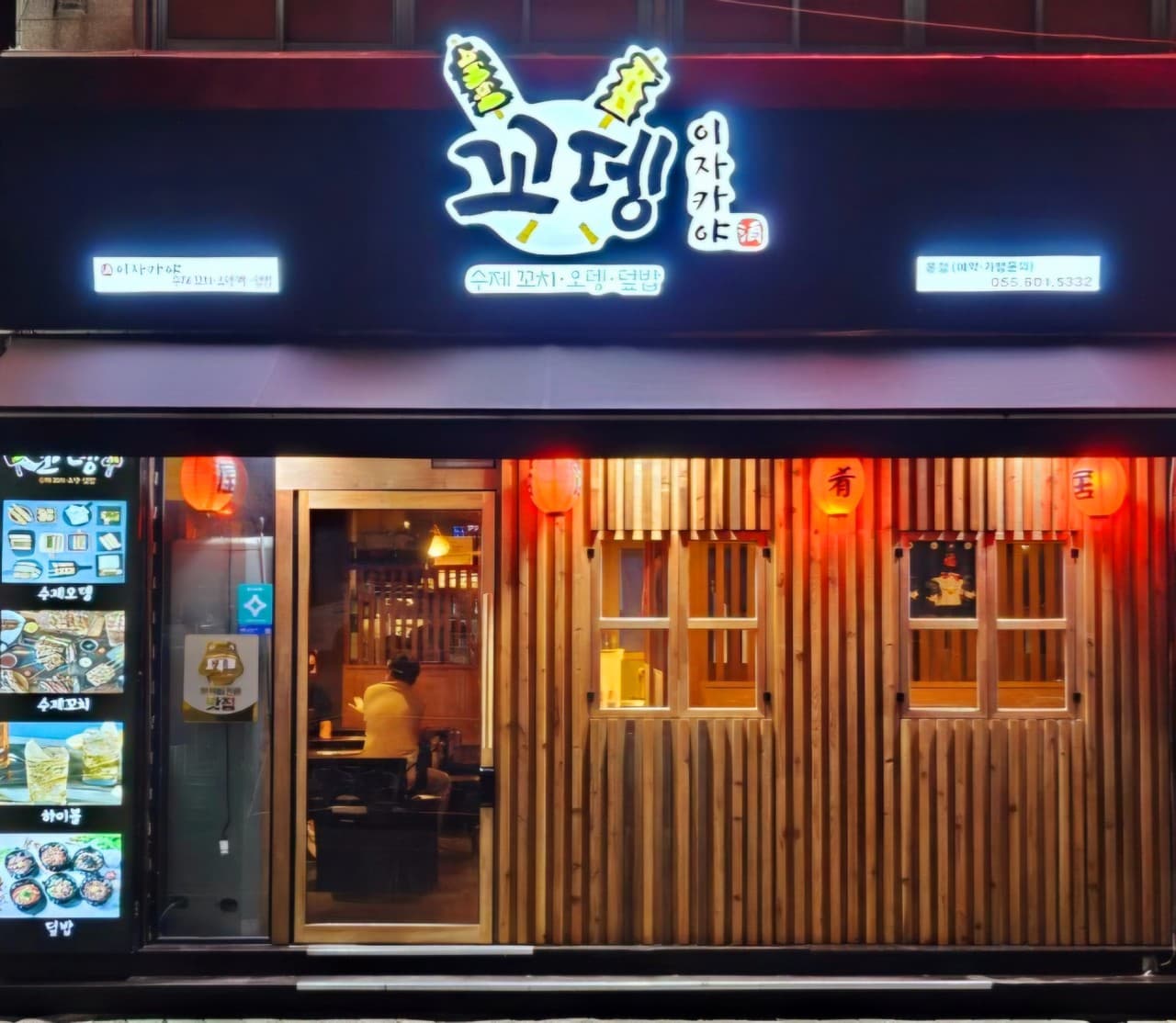 경상남도 창원시 혼술 이자카야 꼬뎅 - 붉은 제등 조명과 나무 외벽 너머 다찌석에 앉은 1인 손님, 수제 꼬치와 오뎅 메뉴판이 보이는 매장 정면