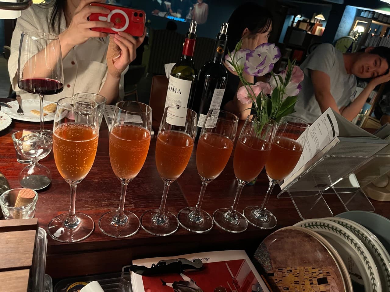 경상남도 진주시 바 사건의장소 다찌석. 전구색 조명 아래 탄산 기포가 보이는 6잔의 로제 와인과 와인병, 책이 놓인 1인 혼술 공간.