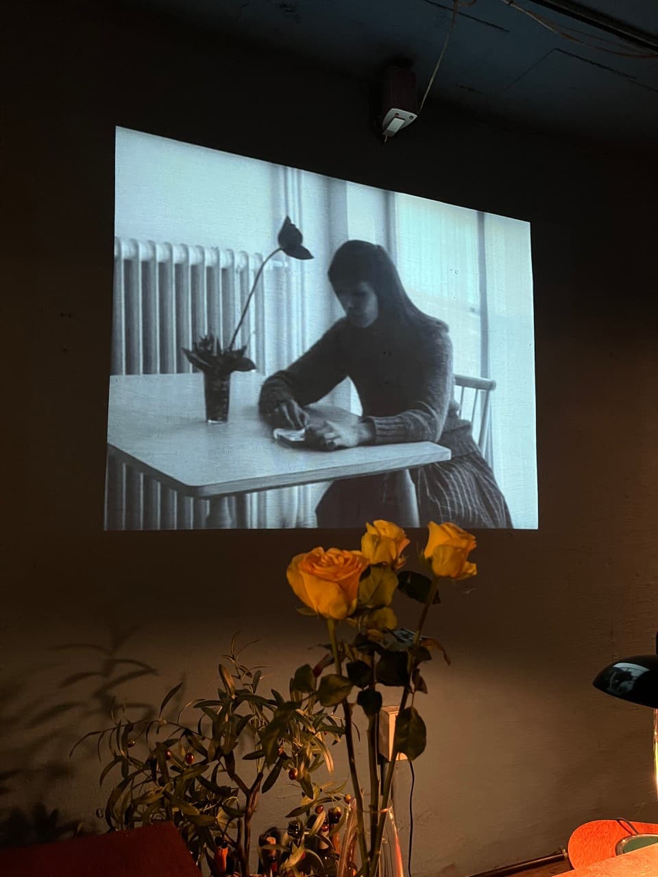 경상남도 진주시 사건의장소 바(Bar)의 벽면 흑백 영화 영사와 노란 장미가 놓인 1인 다찌석. 어두운 조명 아래 홀로 술을 마시는 혼술 상황을 사실적으로 묘사함.