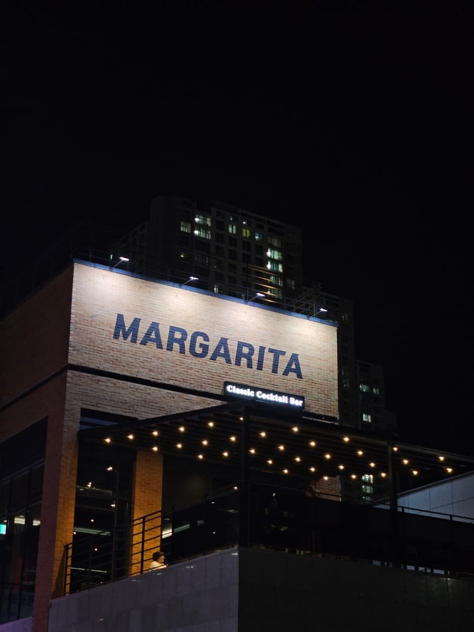 경상남도 진주시 마가리타 진주 바(Bar) 벽돌 건물 외관. 상단 간판 조명과 테라스 아래 배치된 노란 전구들이 어두운 밤을 밝히는 혼술 공간의 모습.