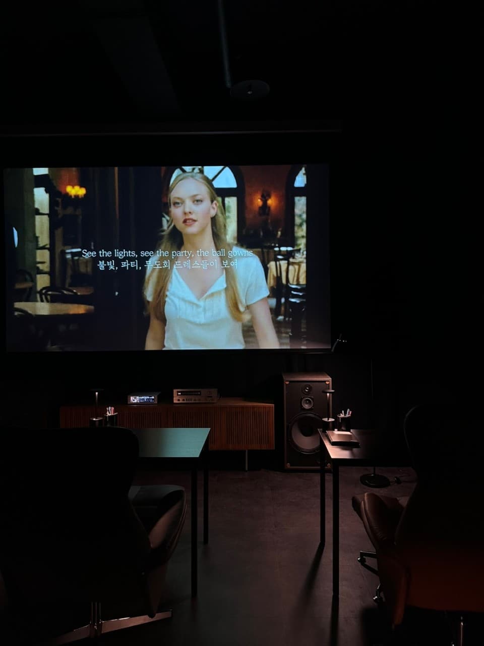 경상남도 진주시 와인 바 스몰그룹 진주 - 영화 상영 스크린과 어두운 조명 아래 1인석 다찌 테이블. 대형 스피커가 있는 공간에서 와인을 즐기는 혼술 상황